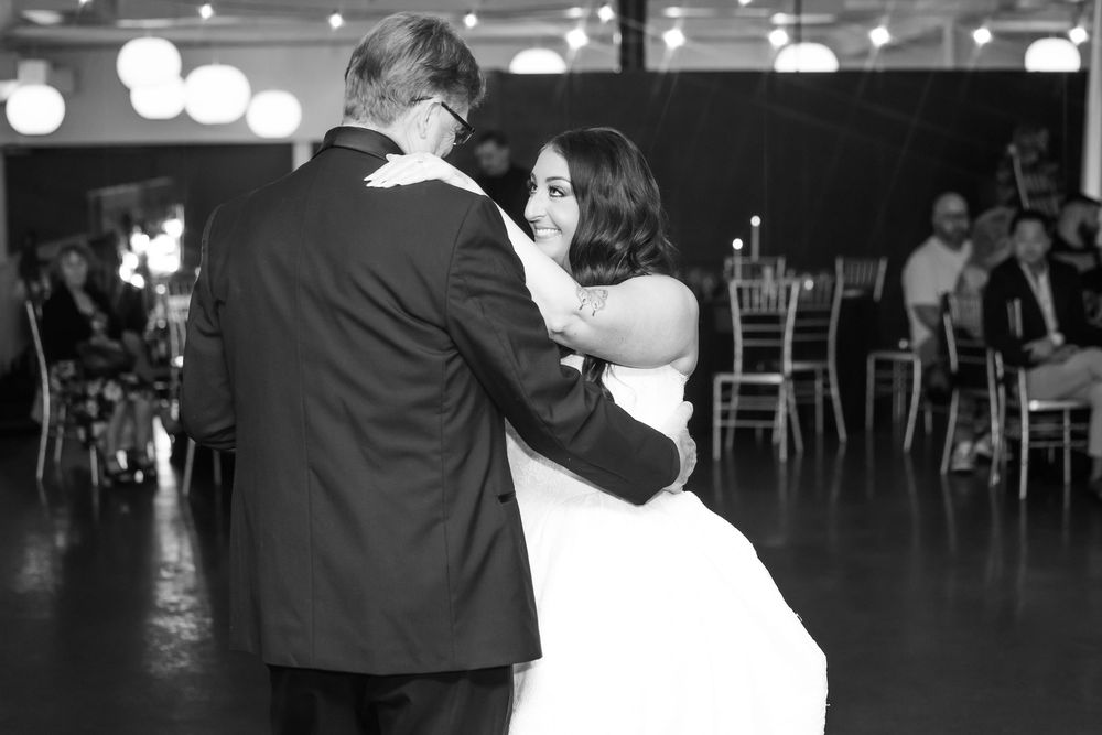 Bride in a white gown shares a first dance with a man in a dark suit at a wedding reception while guests watch from nearby tables.