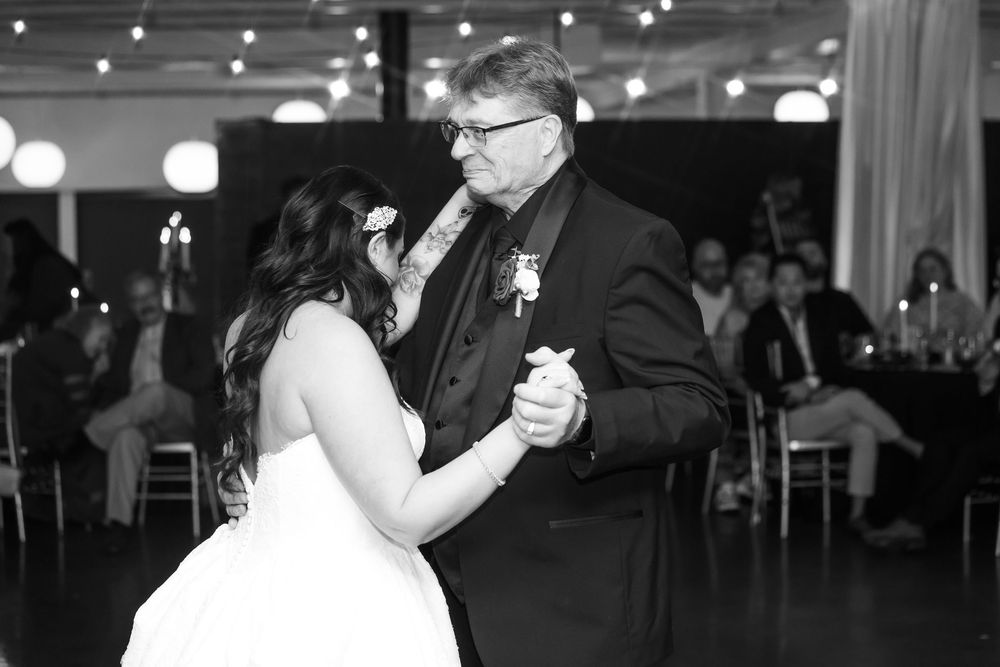 Bride in a white gown dances with an older man in a dark suit and boutonniere while wedding guests watch from tables in the background.
