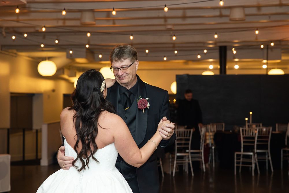 A bride in a white gown dances with a smiling man in glasses and a dark suit at a wedding reception under string lights.