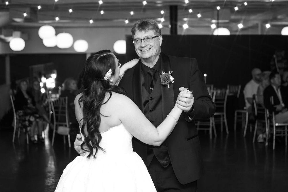 Bride in a white gown dances with a smiling man in glasses and a dark suit with a boutonniere at a wedding reception in black and white.