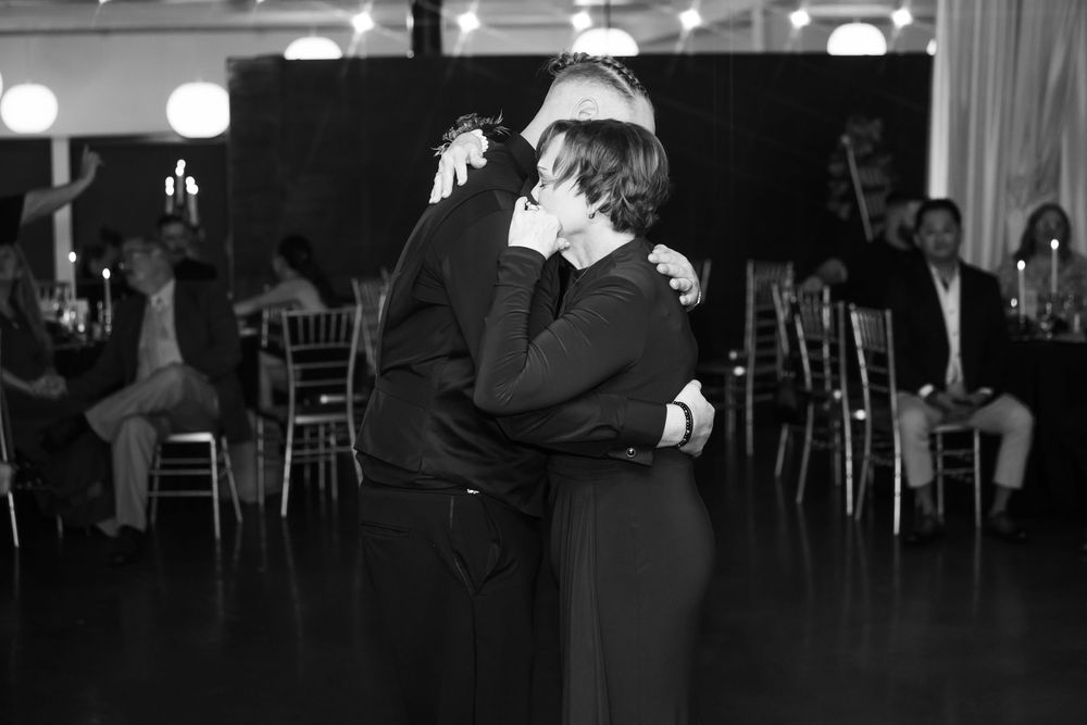 Mother and son share an emotional embrace while slow dancing at a wedding reception, with guests seated at tables in the background.