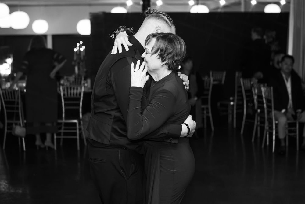 Two people in dark formal attire share a heartfelt embrace on a dance floor at a wedding reception, in black and white.