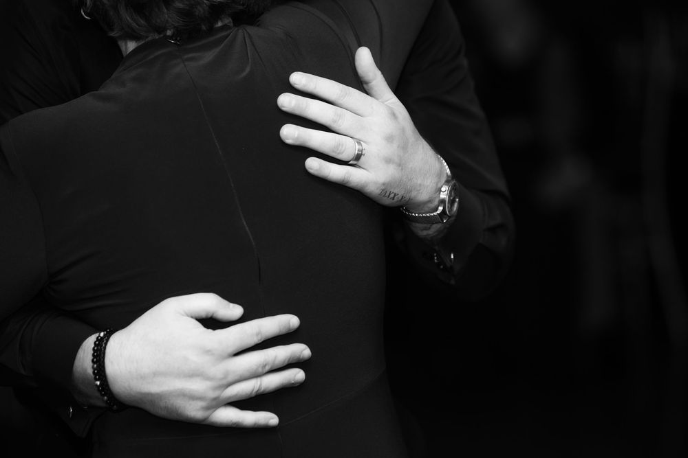 Black and white close-up of two people in a warm embrace, hands resting on dark clothing, with a ring and bracelets visible.