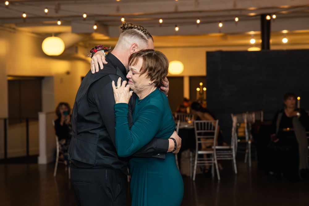 Mother and son share an emotional embrace while dancing at a wedding reception in a warmly lit banquet hall.
