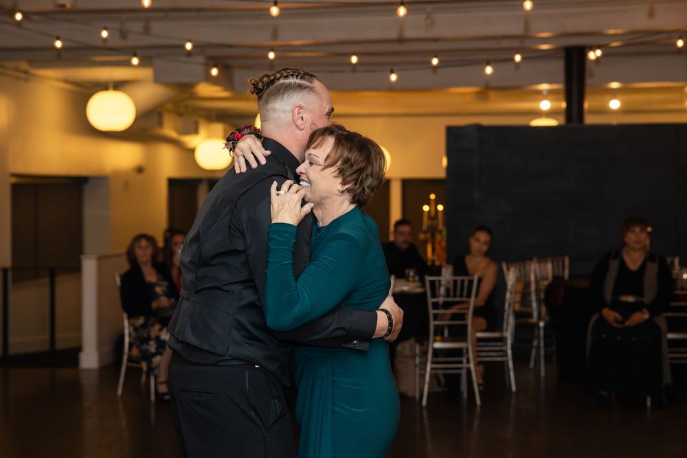 A man in a black shirt and a woman in a green dress share an emotional embrace on the dance floor at an indoor wedding reception.