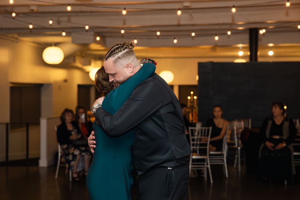 Man in black jacket embraces woman in teal dress during a dance at an indoor wedding reception with guests seated under string lights.