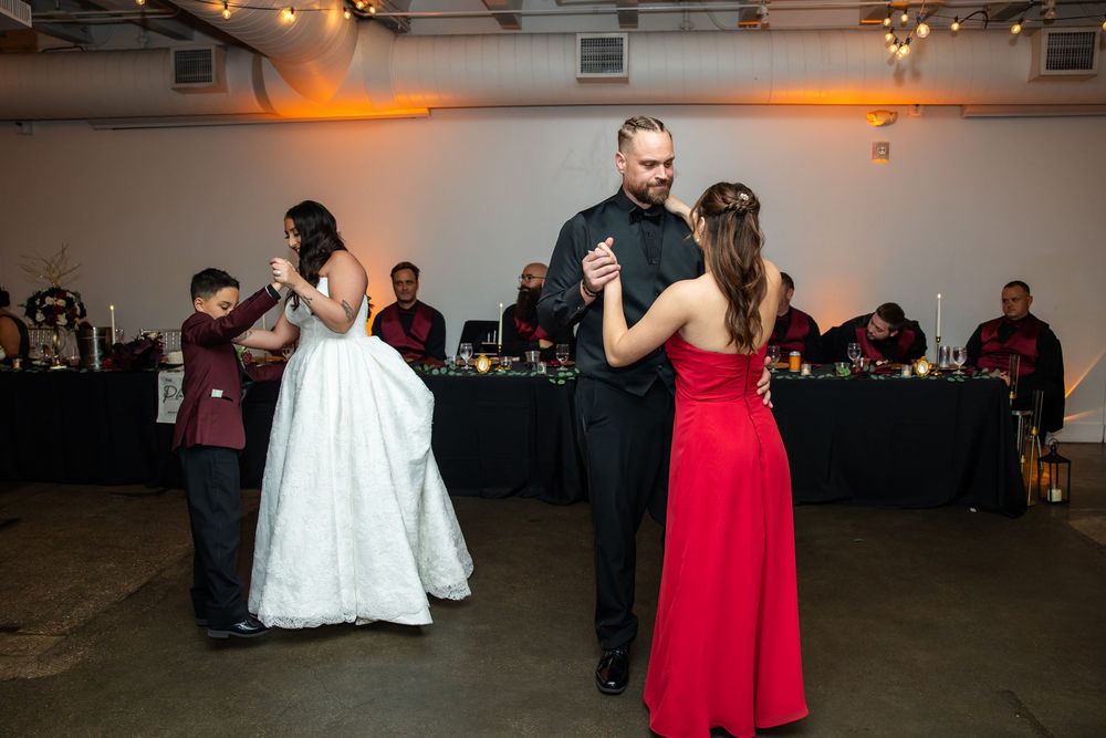 A bride dances with a boy while a groom in black dances with a woman in a red gown at a wedding reception.