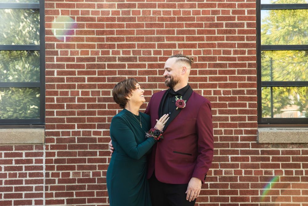 Smiling couple embracing in front of a red brick wall, one in a green dress and the other in a burgundy suit with a boutonniere.