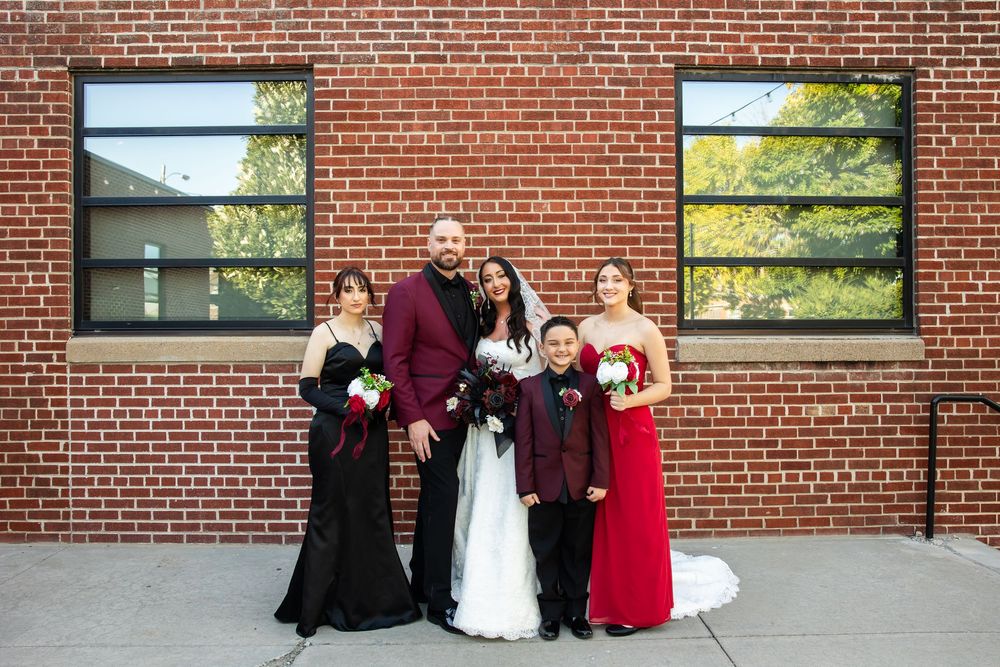 Bride in white gown and groom in burgundy suit pose with a young boy and two women in black and red dresses outside a red brick building.