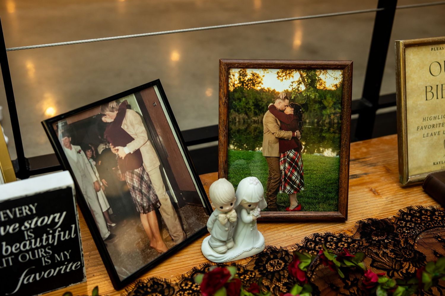 Two framed couple portraits and a small porcelain figurine on a lace table runner at a wedding reception display with a love quote sign.