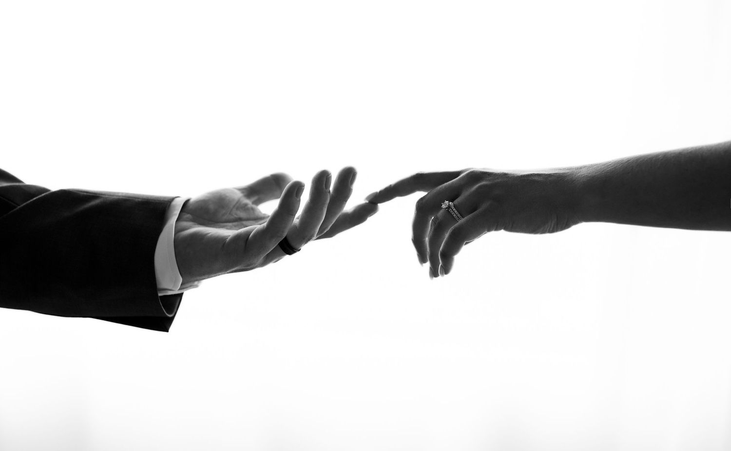 Black and white photo of a man's hand in a suit sleeve reaching toward a woman's outstretched hand against a bright white background.