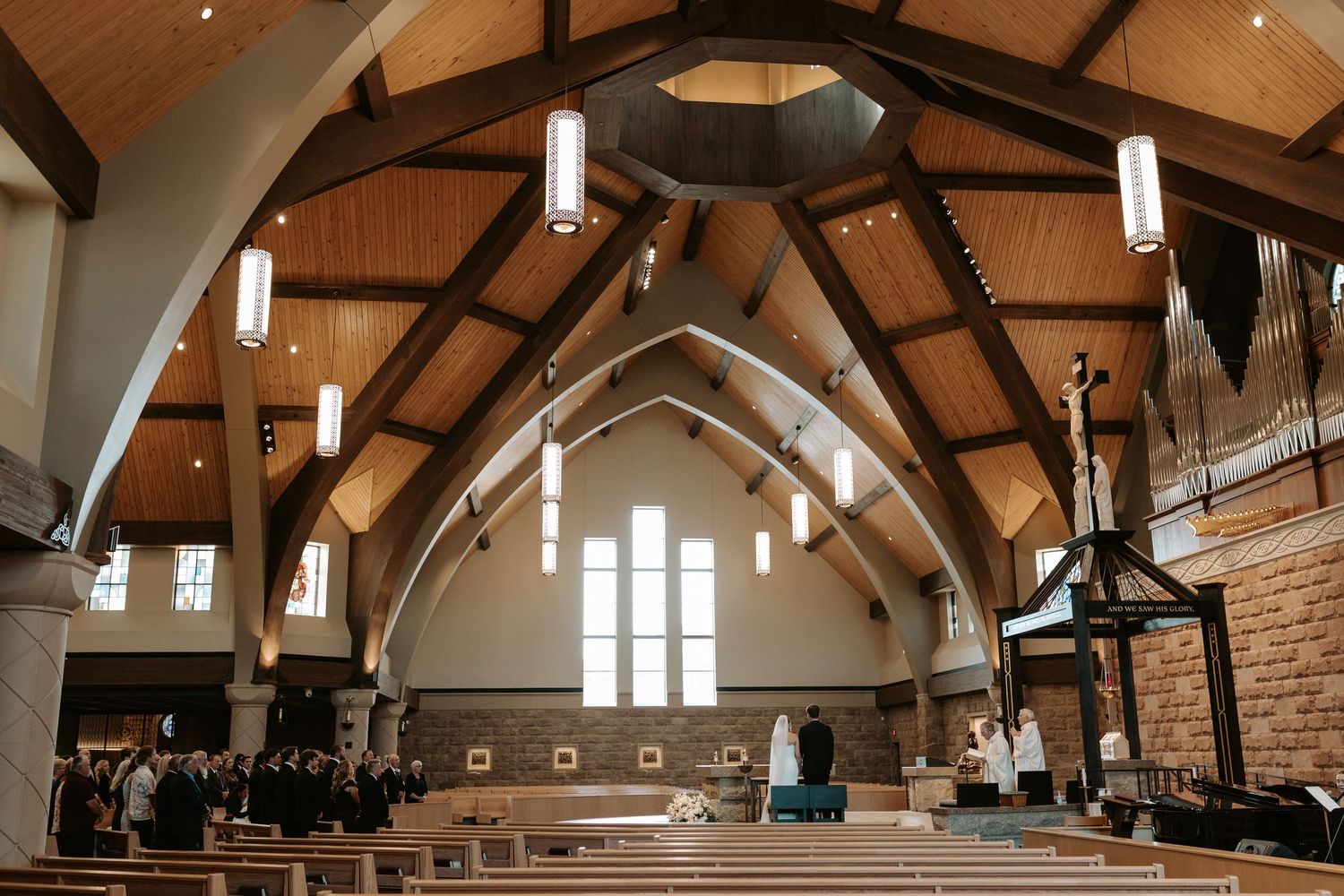 Congregation gathered in a modern church sanctuary with vaulted wood-beam ceiling, pendant lights, stone walls, pews, and tall windows.