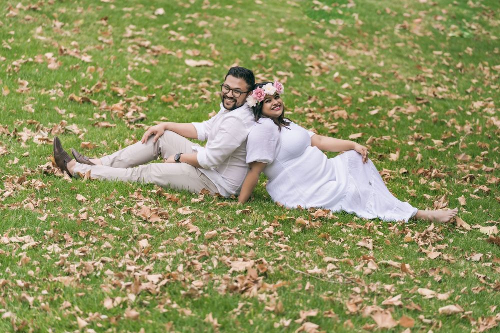 Couple in white outfits sitting back to back and smiling on a grassy lawn covered in fallen autumn leaves.