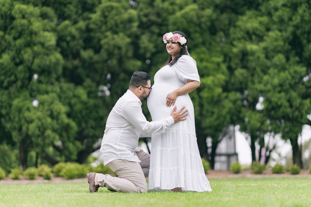 Kneeling man in a light shirt and khakis touches the belly of a pregnant woman in a white dress and floral crown on a park lawn.