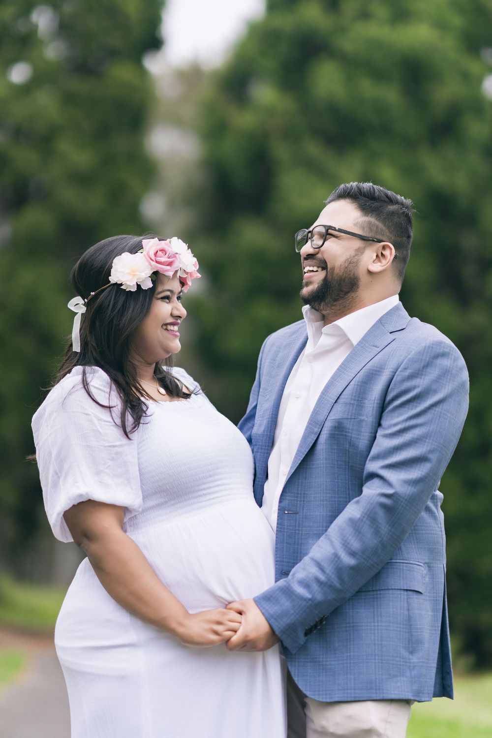 Smiling pregnant woman in a white dress and floral crown holds hands with a laughing bearded man in a blue blazer outdoors.