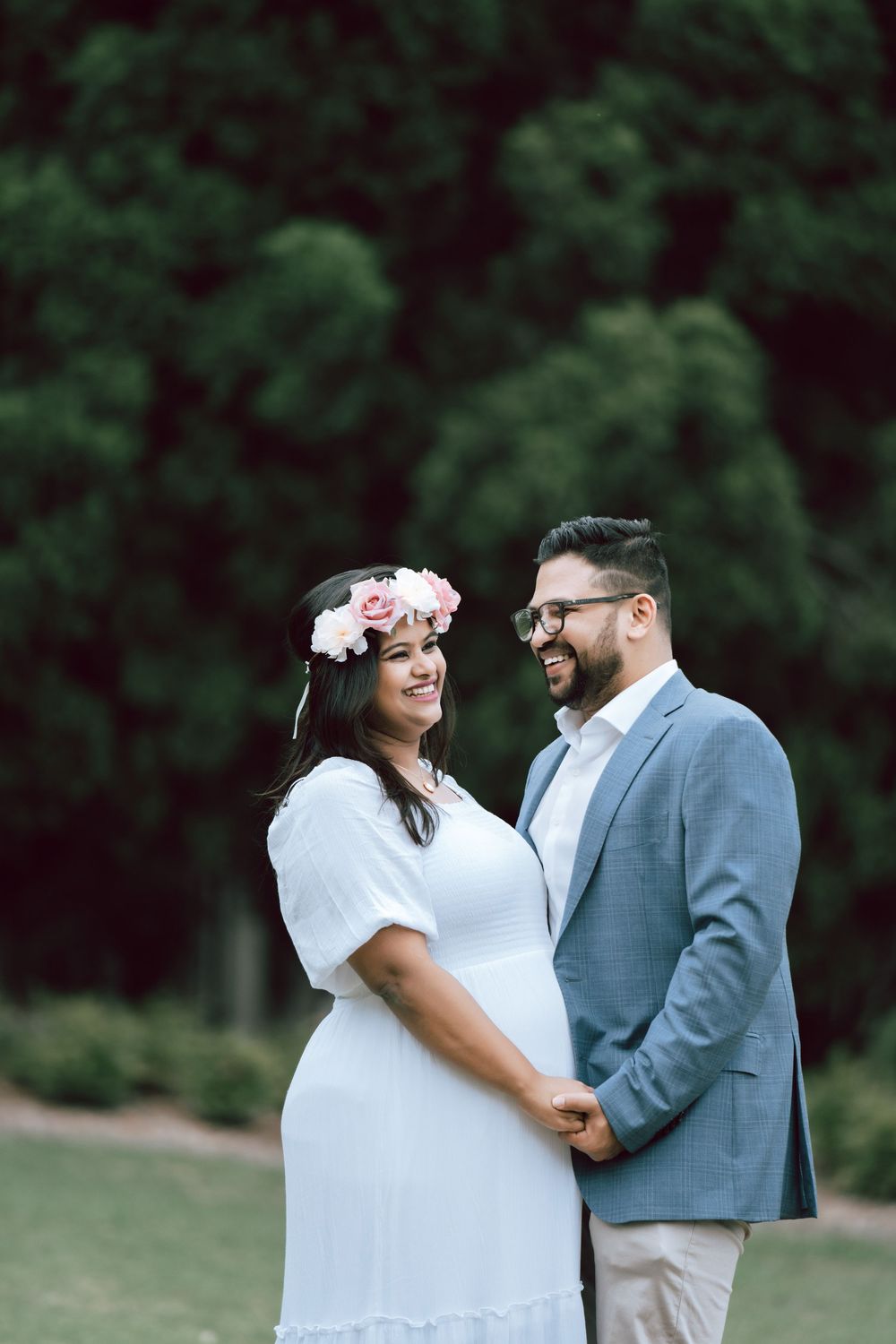 Smiling pregnant woman in a white dress and floral crown holds hands with her bearded husband in a blue blazer in a green park.