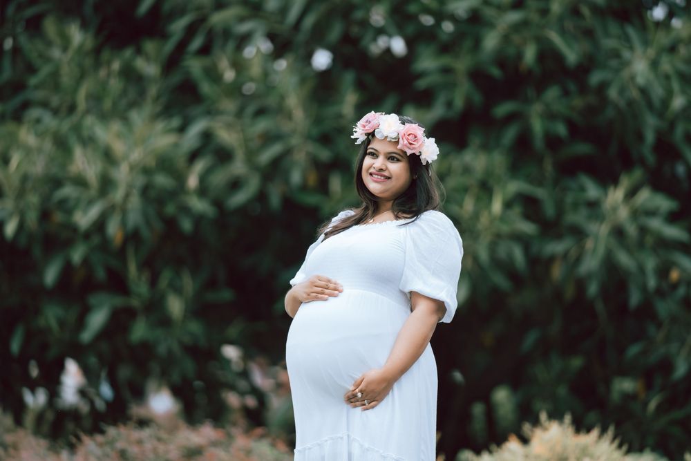 Smiling pregnant woman in a white dress and pink floral crown cradles her baby bump while standing in front of lush green foliage.