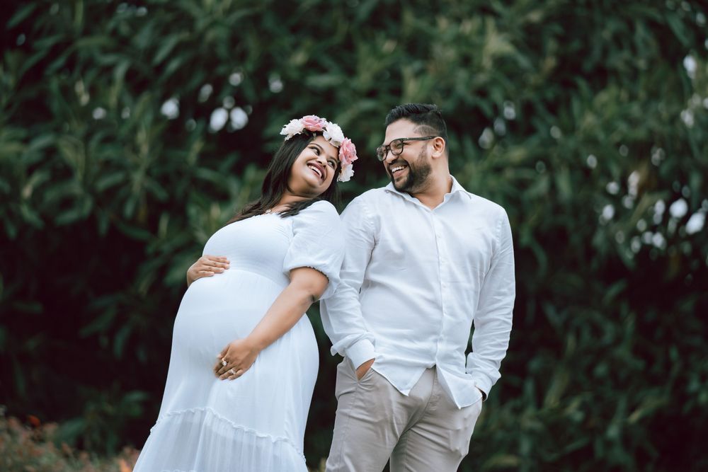 Pregnant woman in a white dress and floral crown smiles as she leans on a bearded man in glasses during an outdoor maternity photoshoot.