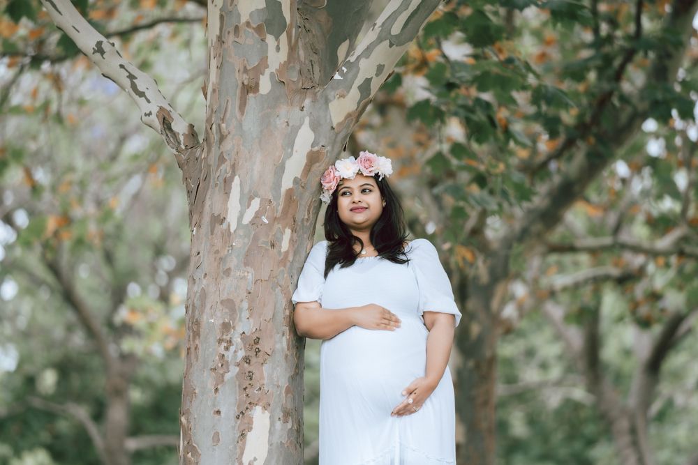 Pregnant woman in a white dress and pink floral crown leaning against a tree trunk in a sunlit park during a maternity photoshoot.