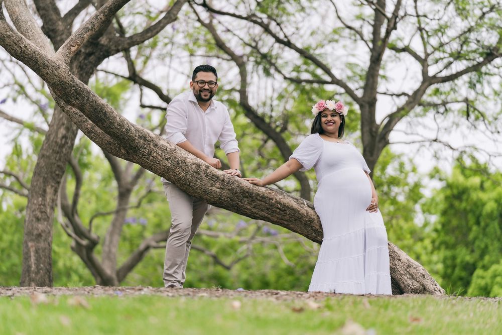 Pregnant woman in a white dress with a floral crown and her bearded husband pose beside a large curved tree branch in a green park.