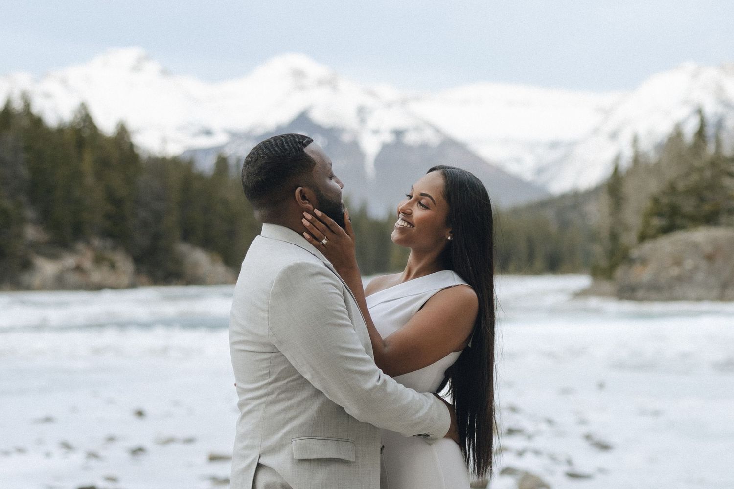 A romantic moment between a couple embracing against a snowy mountain landscape beside a lake.