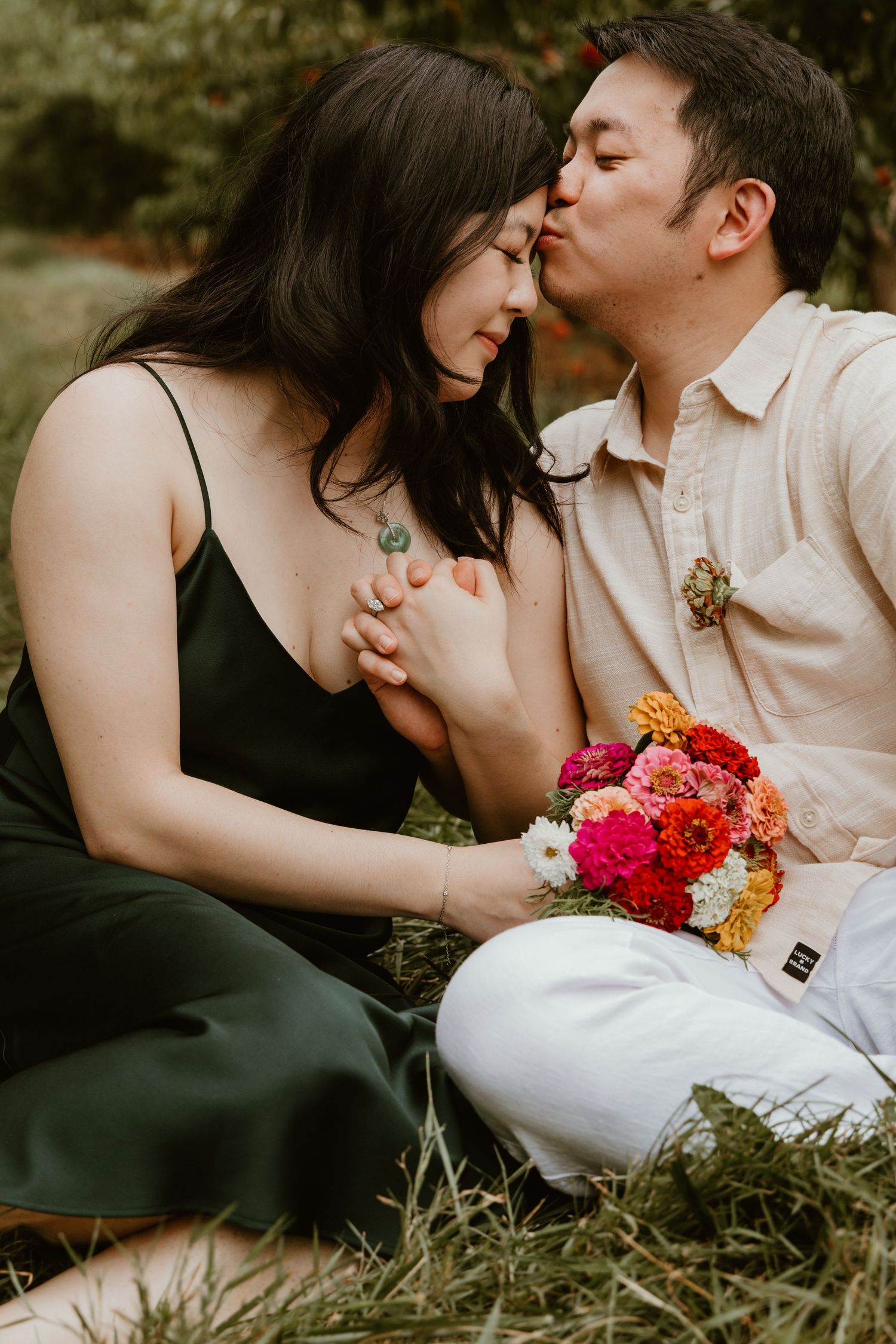 A couple shares a tender kiss while sitting in grass, holding a vibrant bouquet of pink and orange flowers.