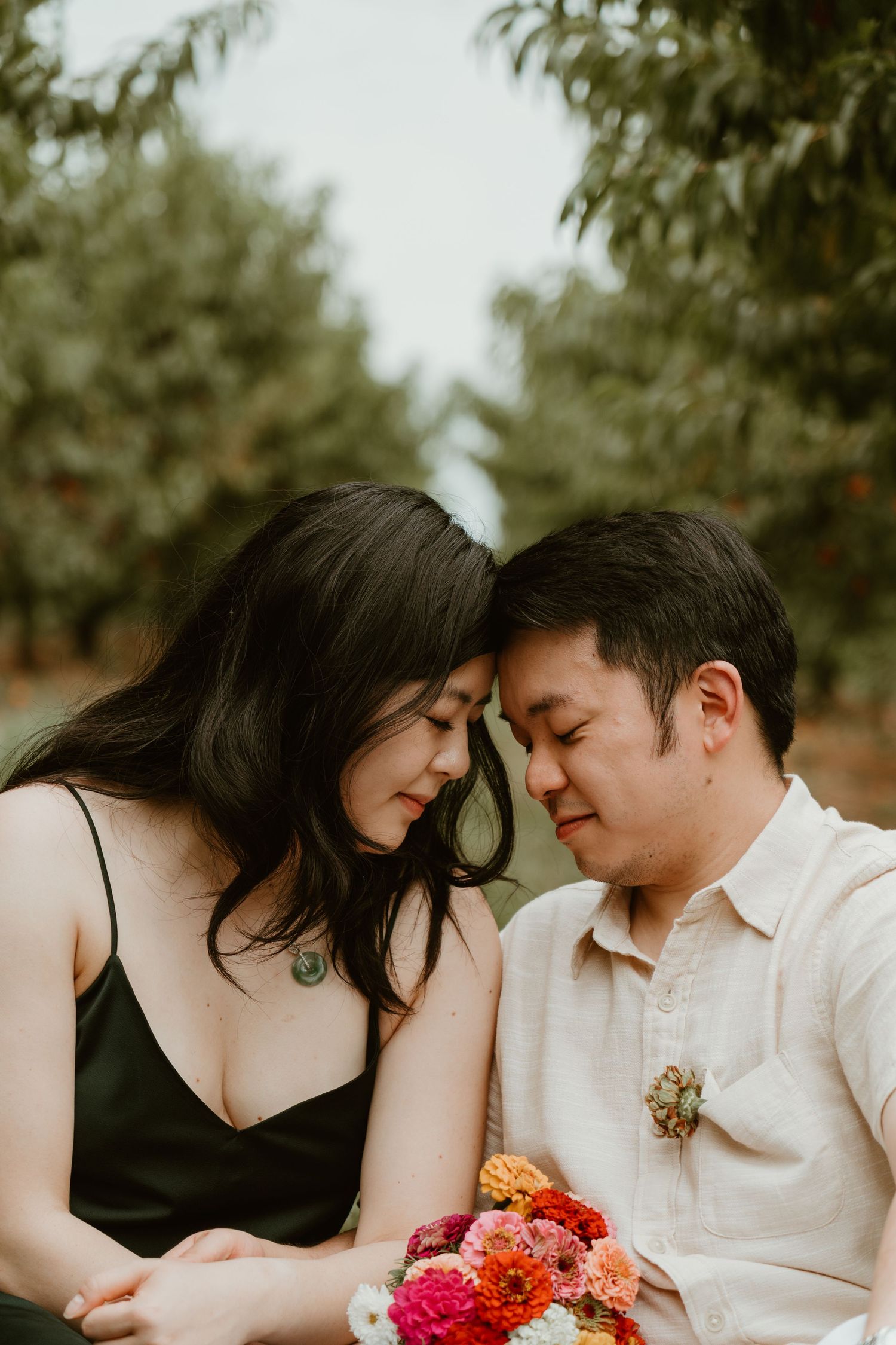 Couple sharing an intimate moment in an orchard, woman in black dress leaning head on mans shoulder holding colorful bouquet.