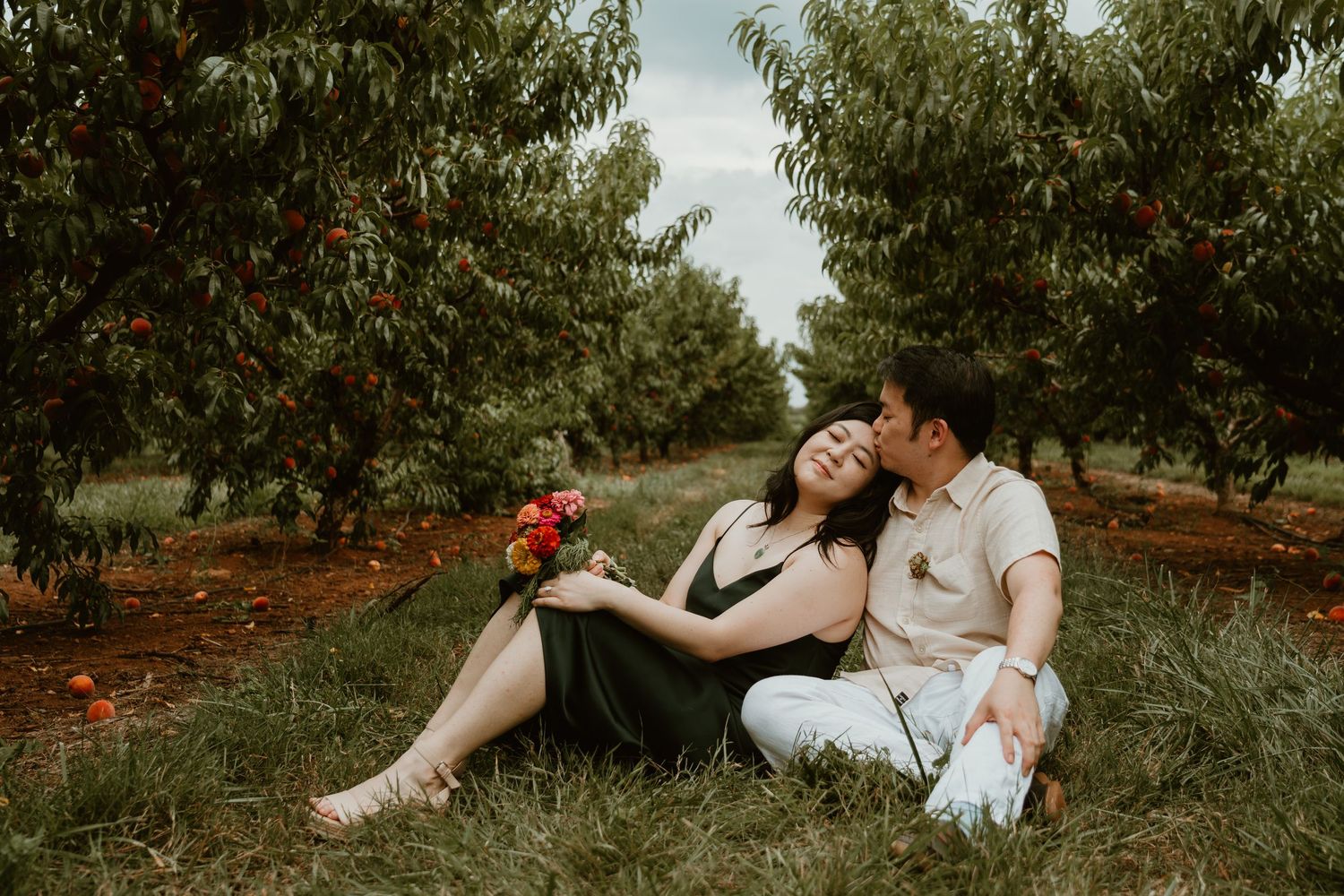 Couple sitting in a peach orchard, woman holding red flowers while man kisses her cheek on green grass.