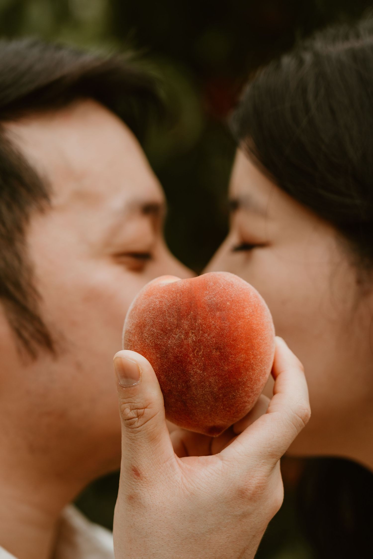 A couple shares a romantic moment kissing behind a fresh ripe peach held between them outdoors.