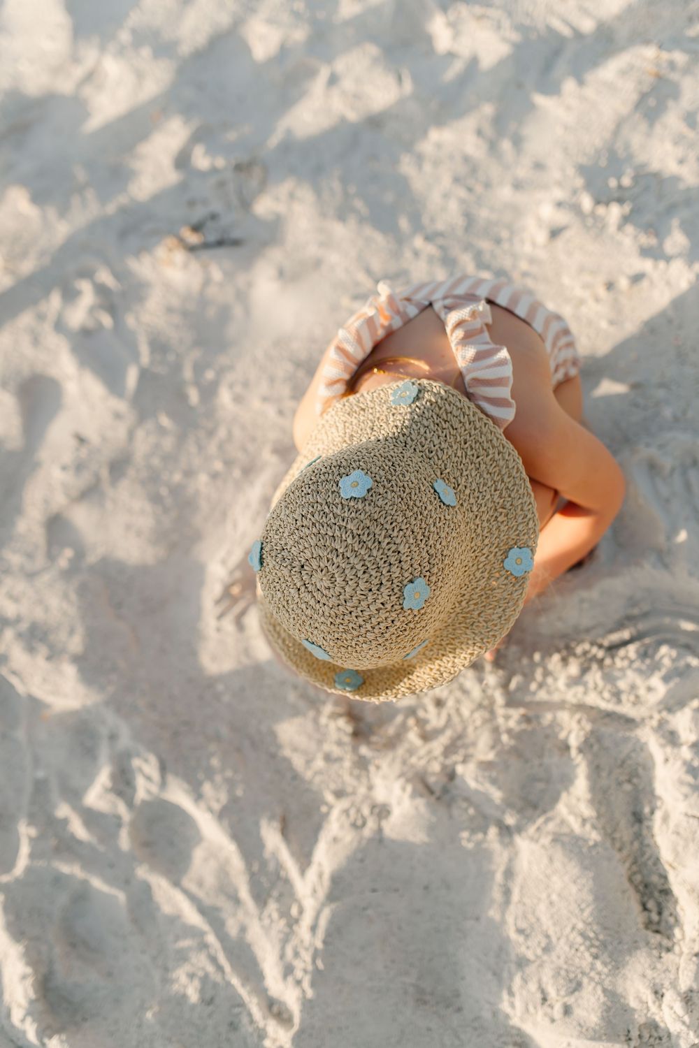 Young child sitting on sandy beach wearing a straw hat with blue hearts and striped swimsuit top.