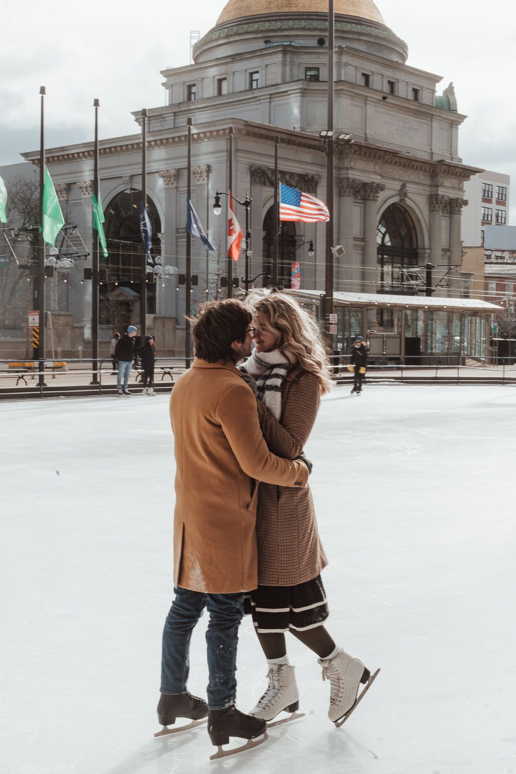 Romantic couple kissing on outdoor ice skating rink in winter, wearing brown coats, with historic building and American flag backdrop.