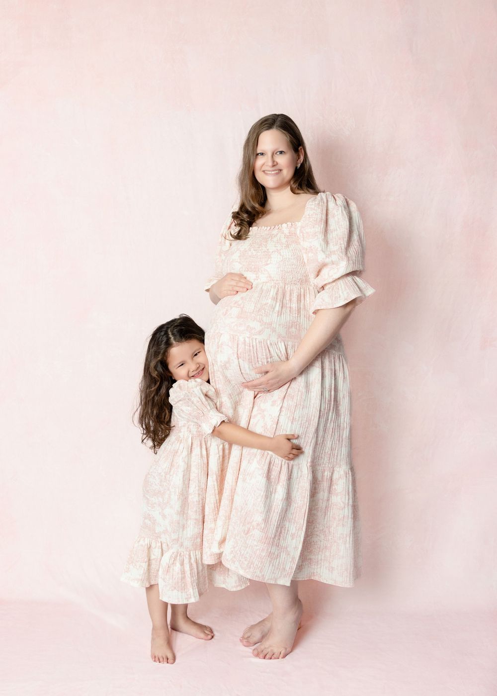 Pregnant woman and young girl in matching floral dresses pose together for a maternity photo session.
