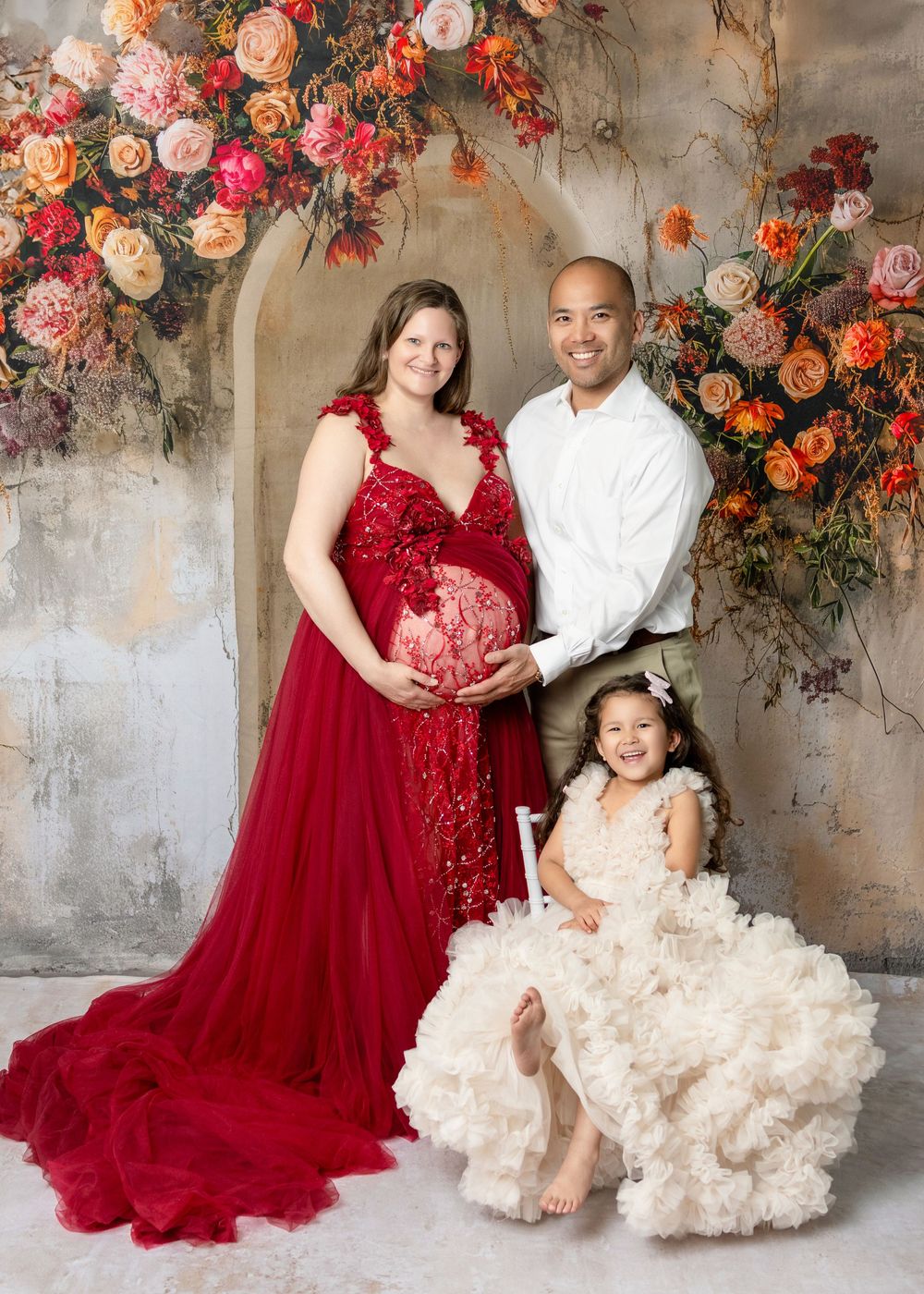 Pregnant woman in red gown poses with husband and daughter before floral arch backdrop in maternity photo.