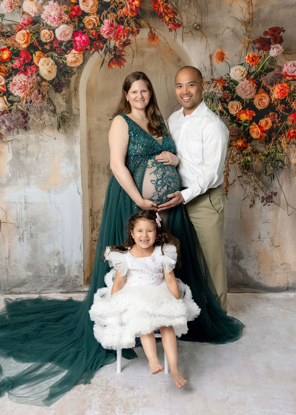 Maternity photo of pregnant woman in green gown, man in white shirt, and young girl in white dress with floral backdrop.