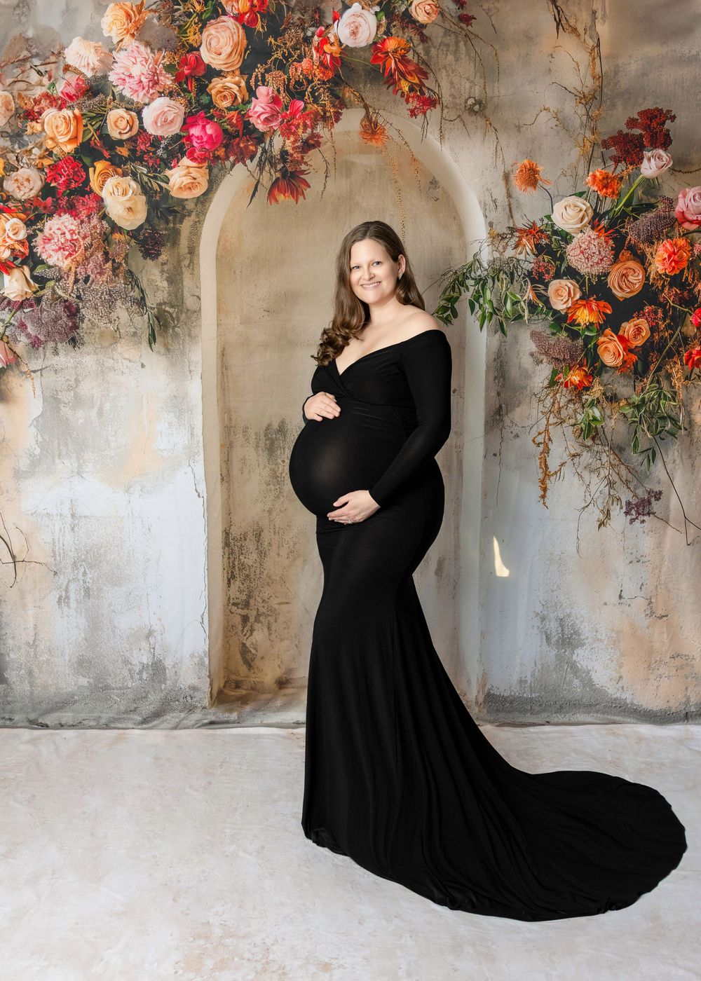 Pregnant woman in elegant black off-shoulder gown poses before floral arch backdrop in professional maternity portrait.