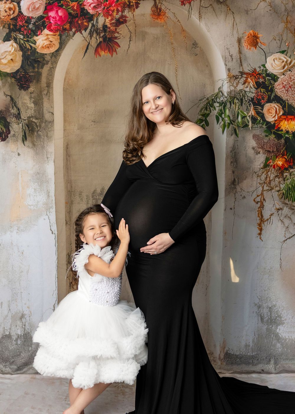 Pregnant woman in black gown poses with young girl in white dress before floral arch backdrop.