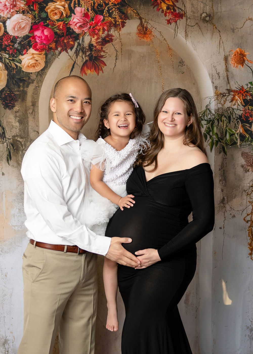 A smiling family maternity photo with a man, young girl, and pregnant woman in black dress before a floral arch backdrop.