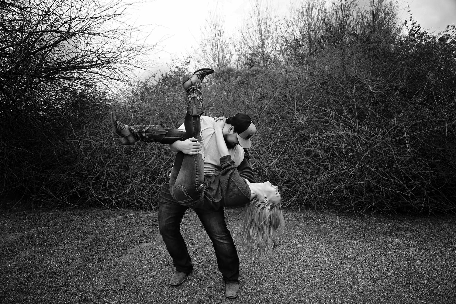 A couple plays in a black and white photo as a man lifts a laughing woman upside down by a dirt path lined with bare bushes.