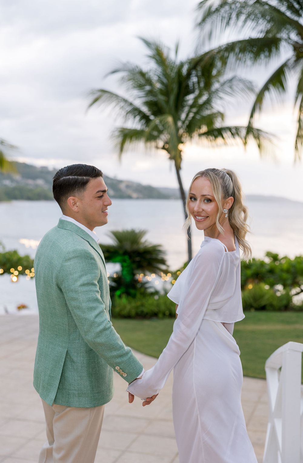 Couple holding hands at tropical beach wedding venue with palm trees and ocean backdrop at sunset.