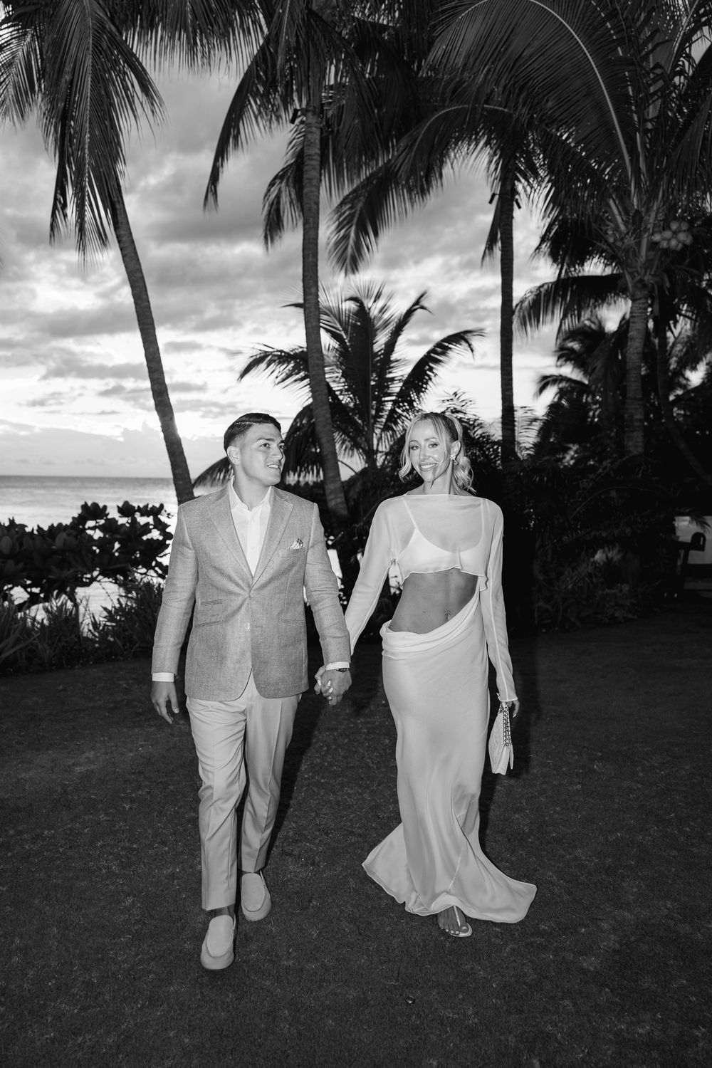 Black and white photo of a couple holding hands walking among palm trees near the ocean, woman in elegant white gown.