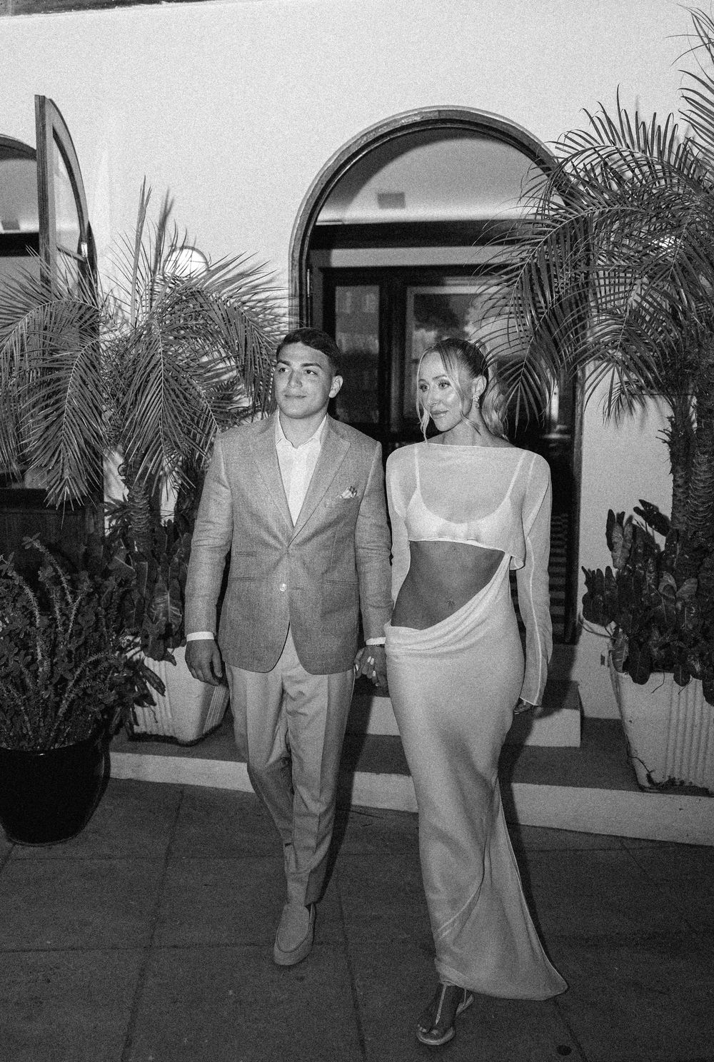 Couple posing outside elegant venue, man in suit and woman in sheer cutout gown, black and white photo.