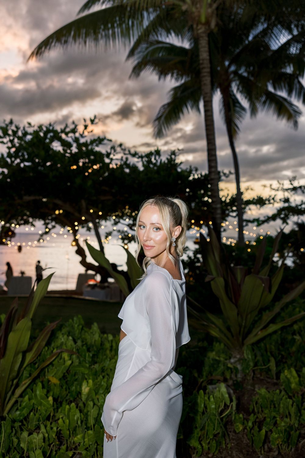 Woman in white dress poses at tropical sunset wedding venue with palm trees and string lights.