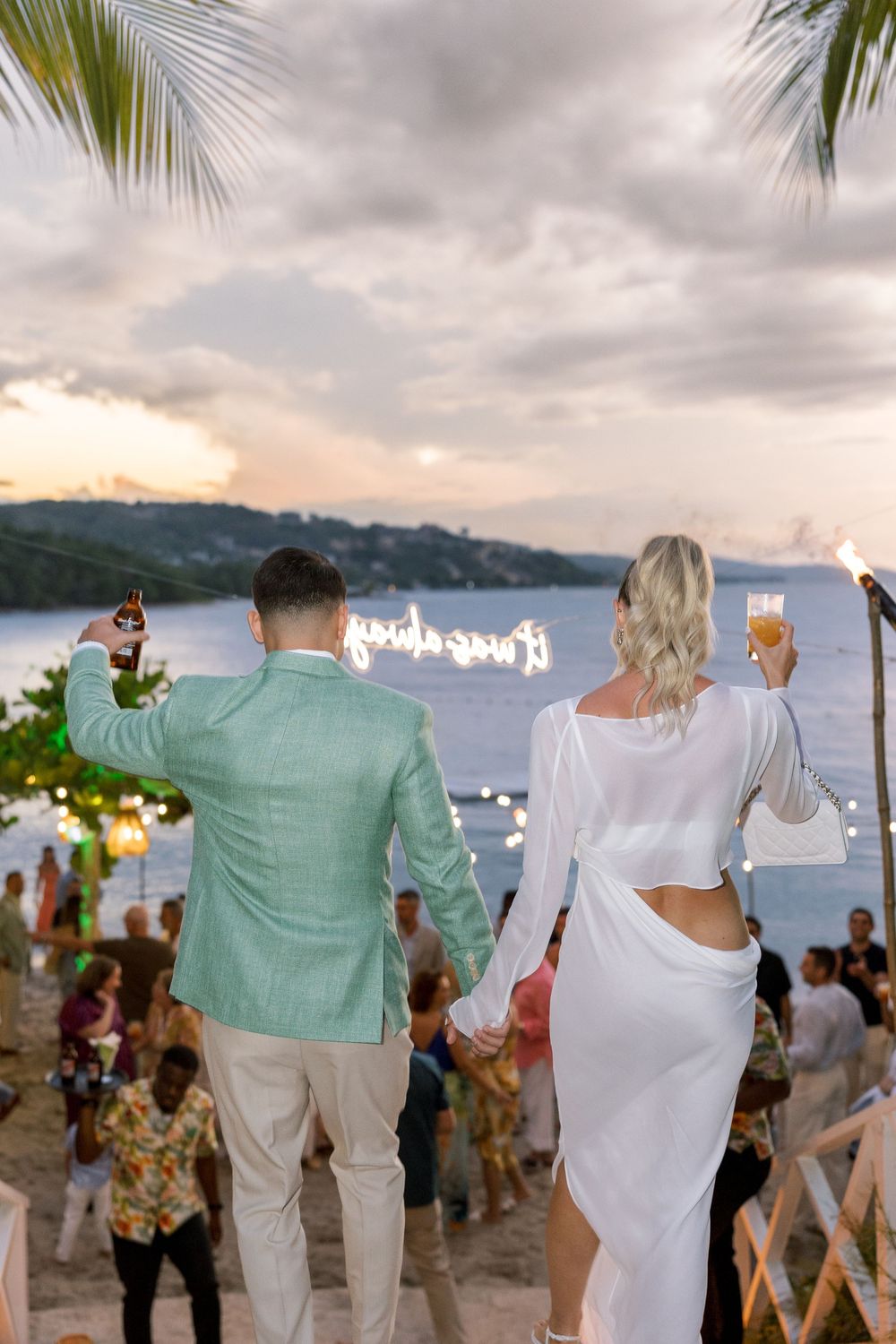 Couple in white and mint green holding champagne at a beach wedding reception at sunset.
