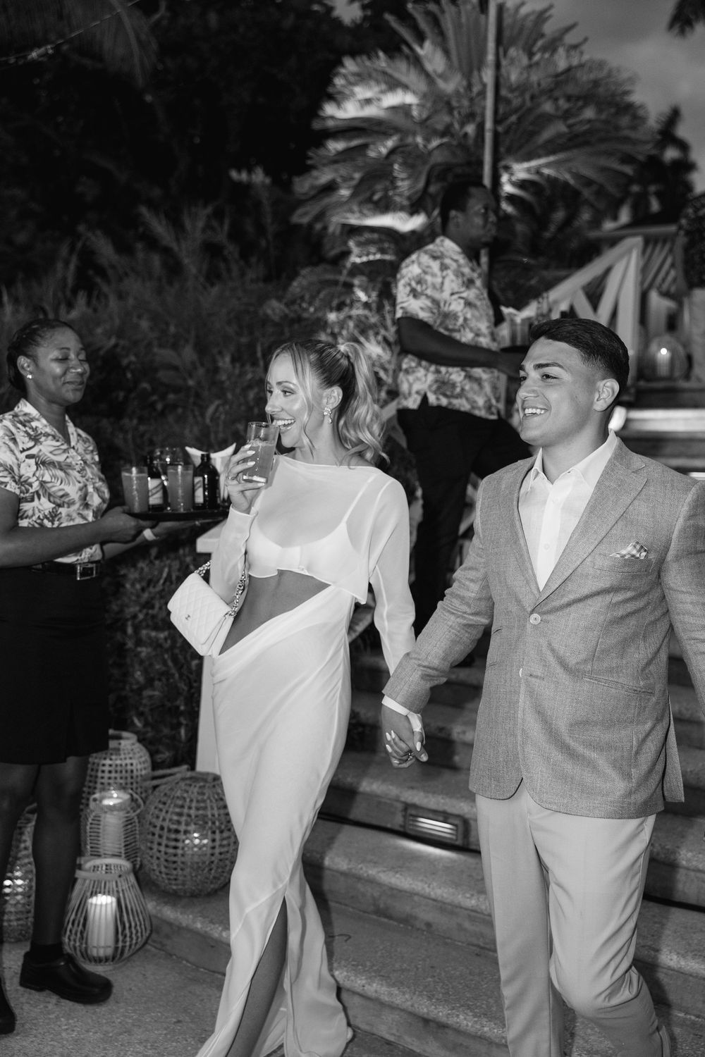Black and white photo of a couple holding hands at an outdoor evening wedding reception near tropical palms.