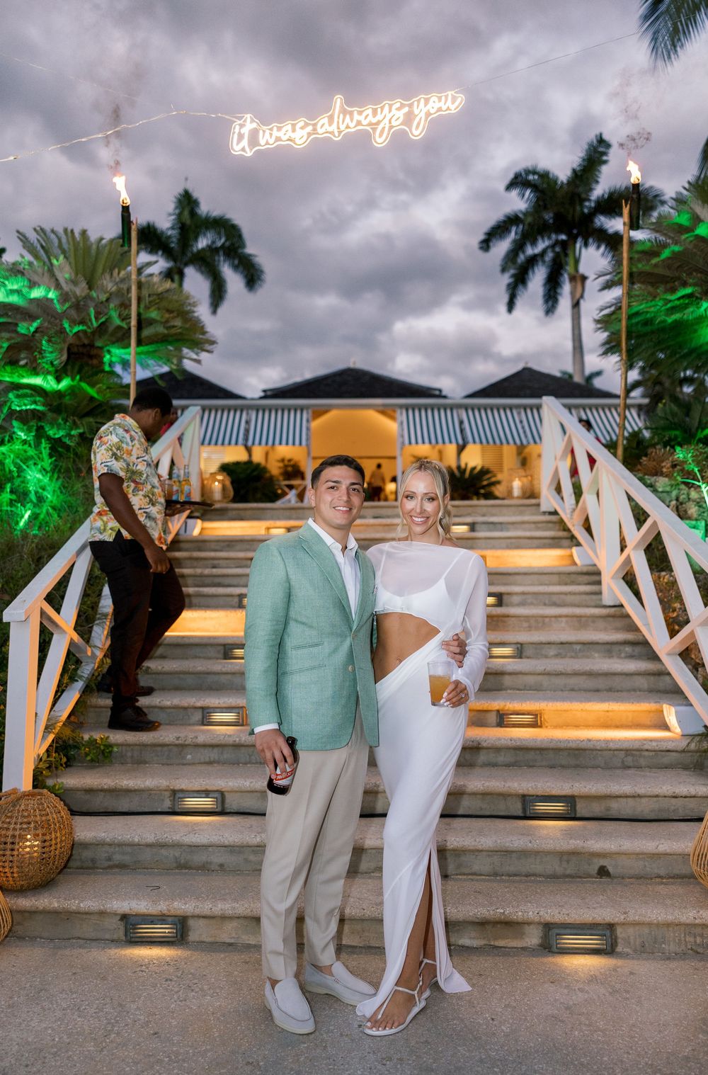 Couple poses on lit stairs at tropical venue with neon sign reading it was always you at dusk.
