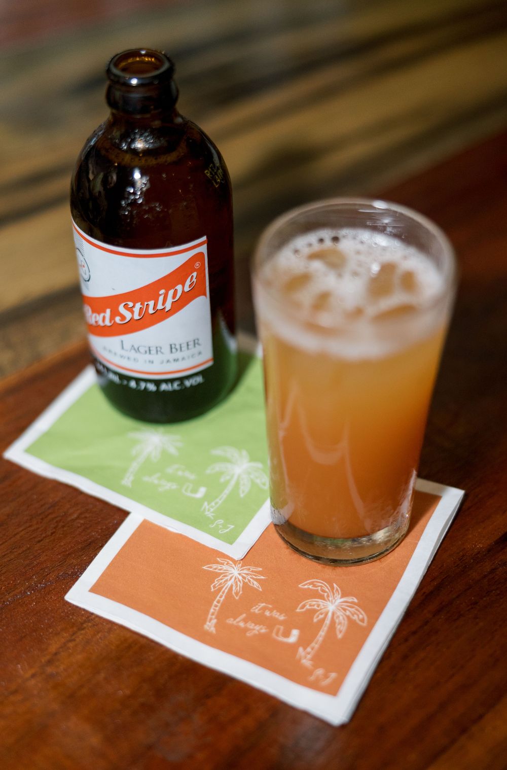 Red Stripe Jamaican lager beer bottle beside a chilled glass of beer on tropical palm tree cocktail napkins.