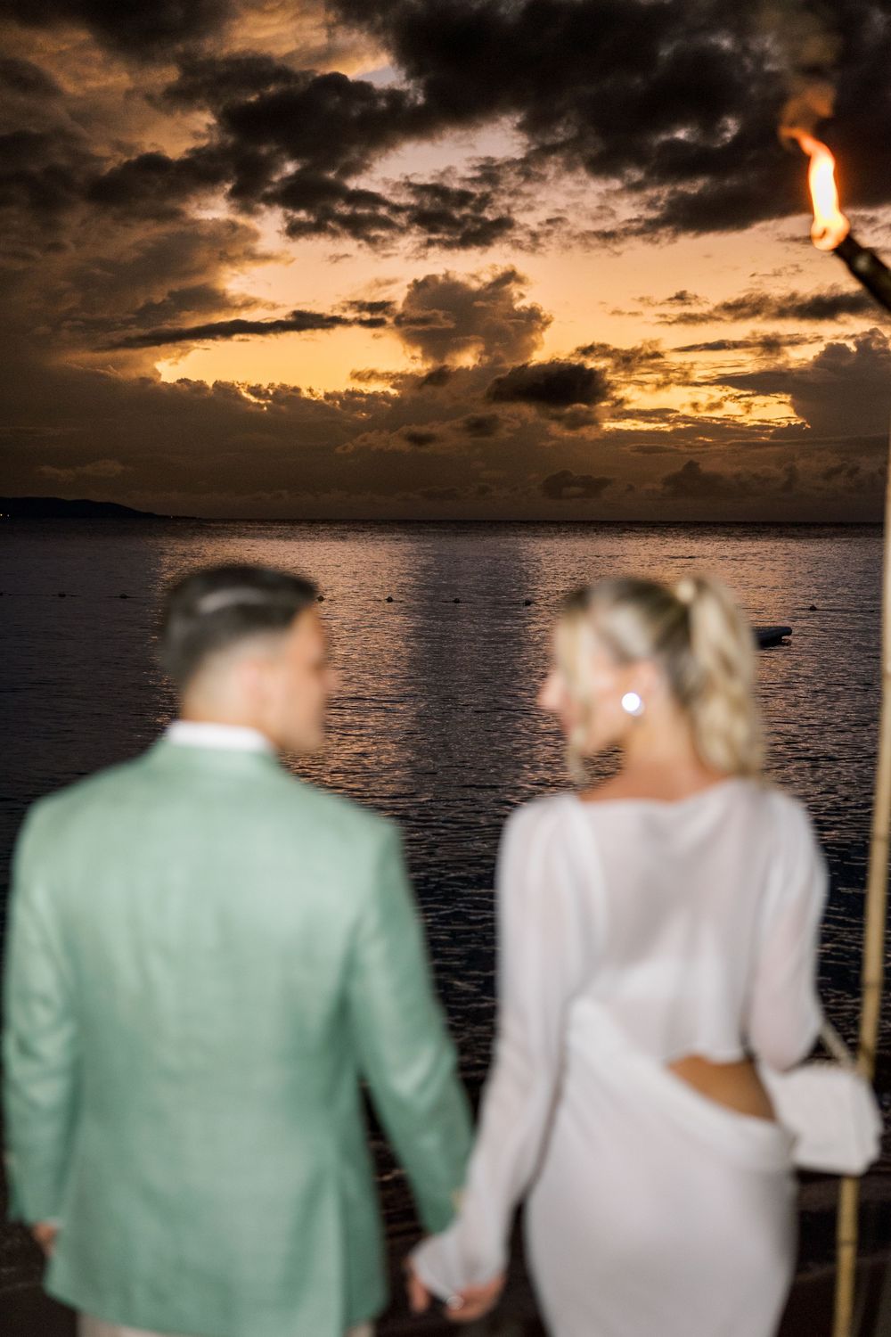 Couple holding hands at sunset by the ocean, man in mint suit and woman in white dress, with a tiki torch glowing above.