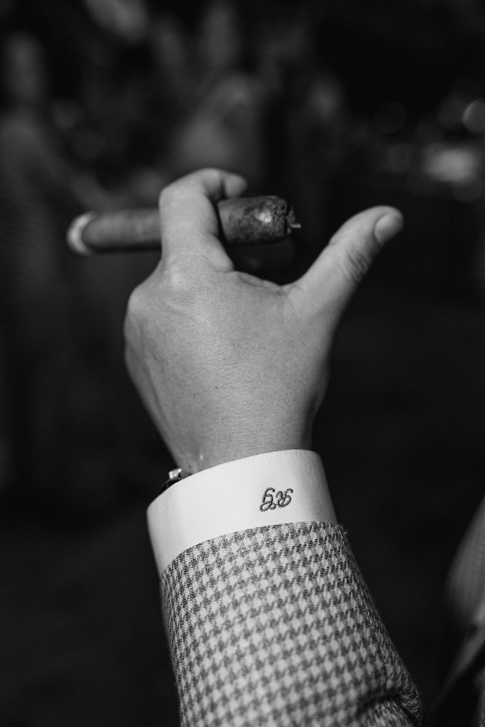 Black and white photo of a man in houndstooth suit holding a cigar, monogrammed shirt cuff visible.