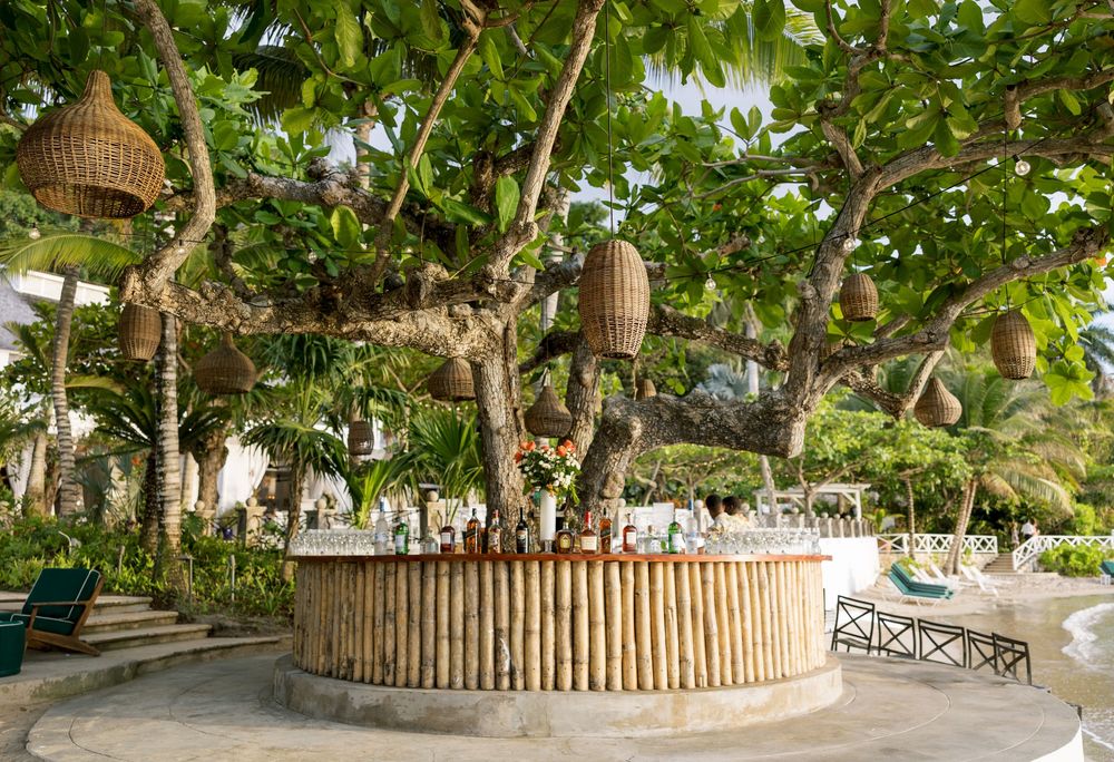 Tropical outdoor bamboo bar under a large tree with hanging wicker lanterns in a lush resort garden.