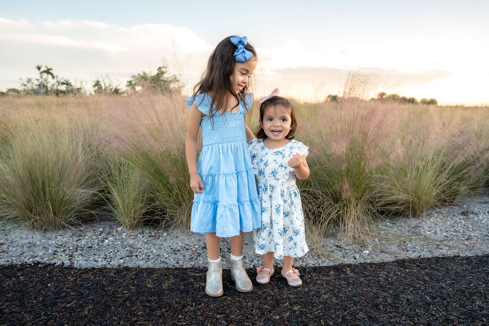 Two young girls in blue dresses pose together outdoors near ornamental grasses at sunset.